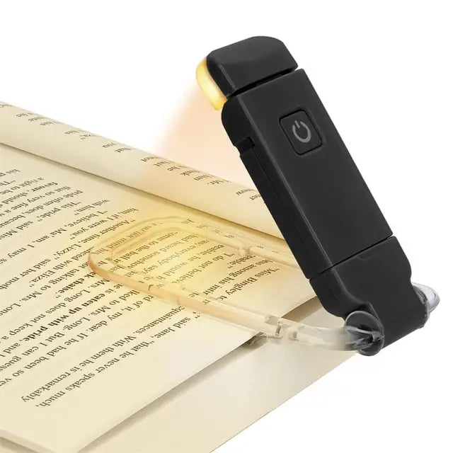 A black clip-on book light with a built-in USB charging port, shown clipped to an open book on a white background.