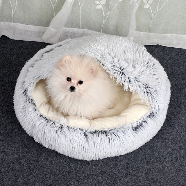 Small white dog in a fluffy gray pet bed on a dark surface with a light gray background